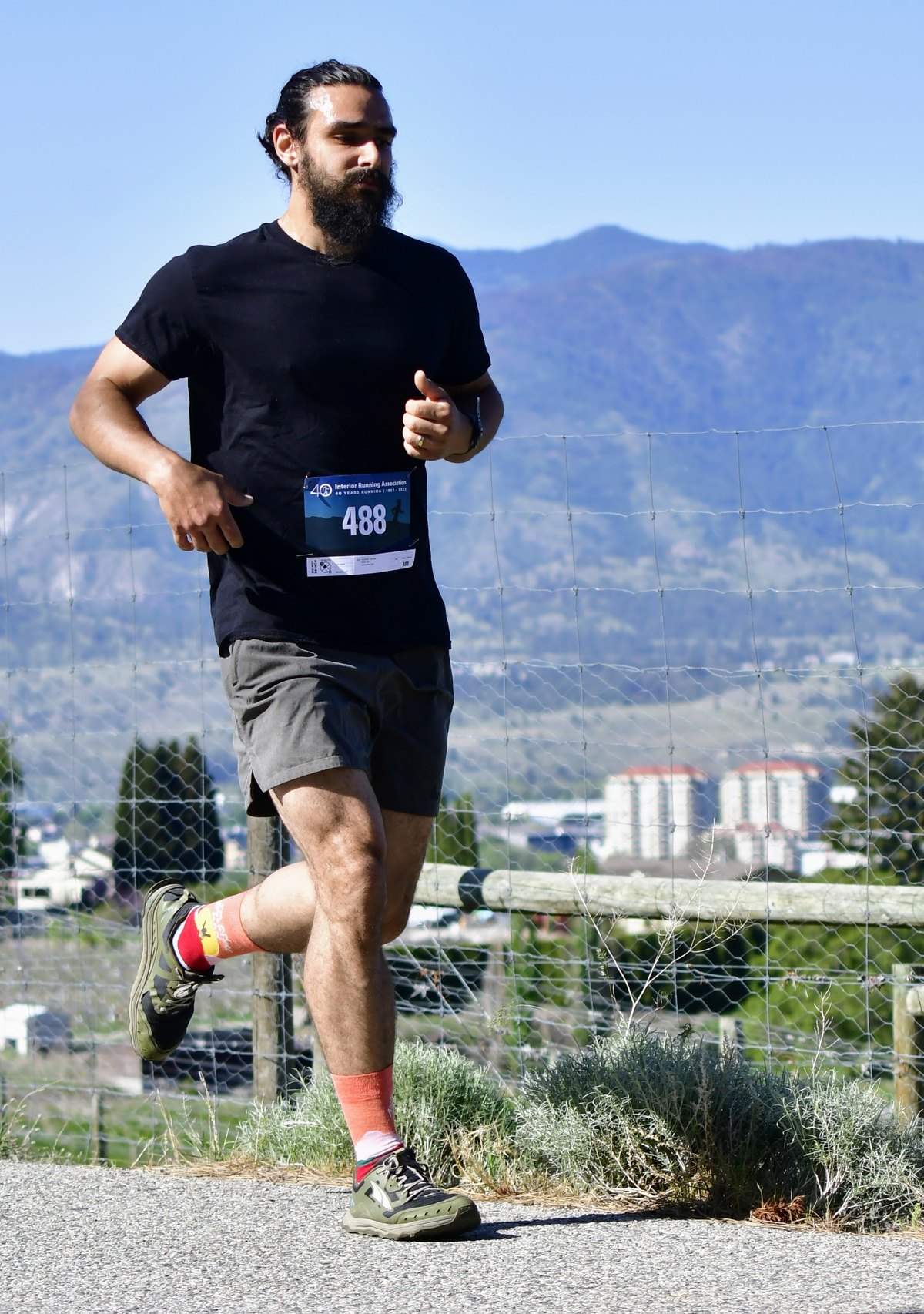 A runner in a black tee and race bib holding form on a sun-baked Okanagan road, mountains behind